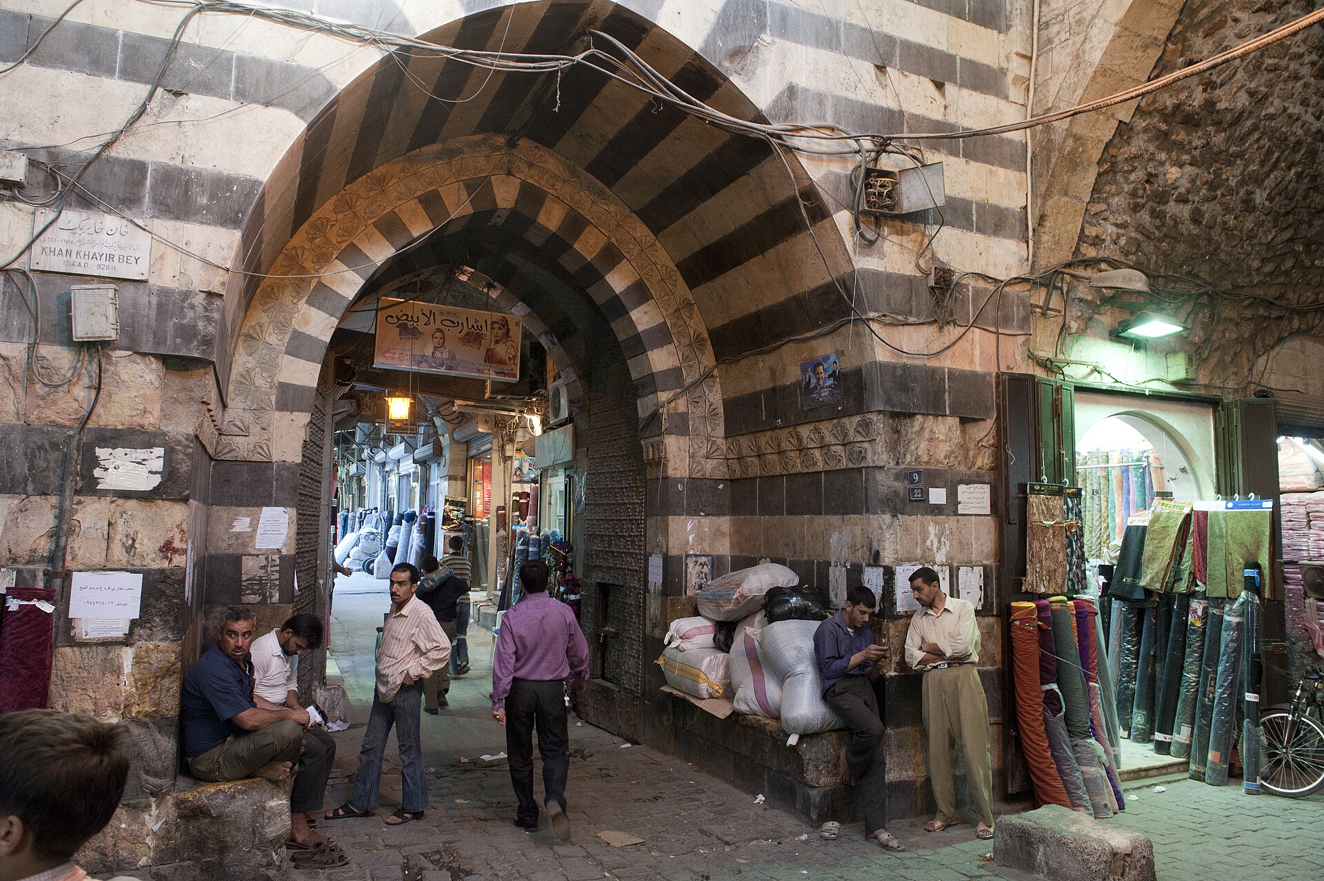 A striped stone archway in an Aleppo souq, with traders and goods gathered beneath it.